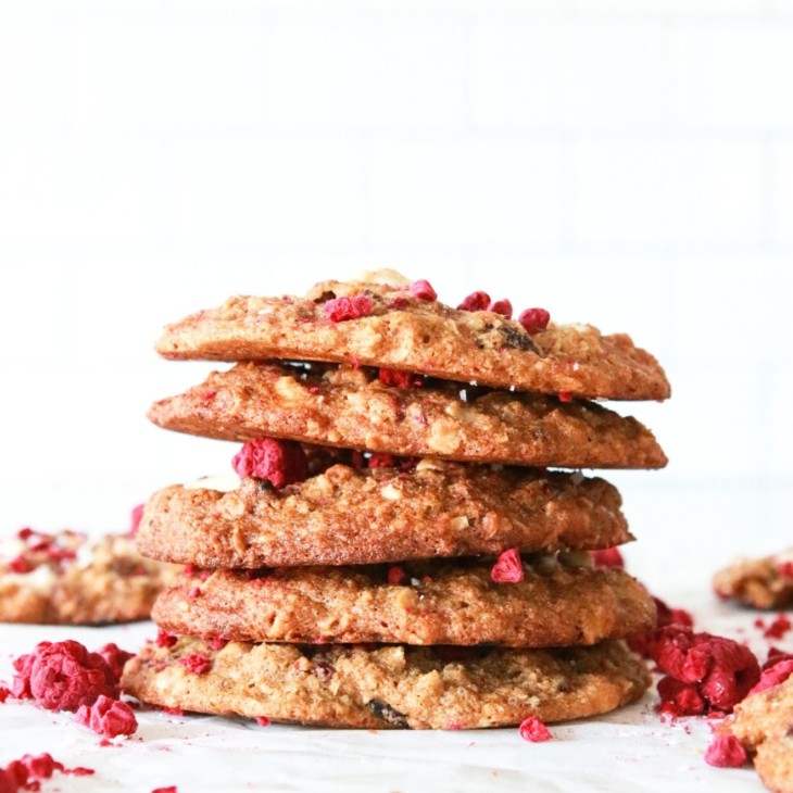 This is a side view of a stack of gluten free cookies. The stack sits on a white piece of parchment paper and a white background. Freeze dried raspberries are around the cookies.