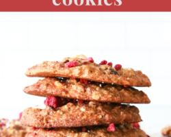 This is a side view of a stack of cookies. The stack sits on a white piece of parchment paper and a white background. Freeze dried raspberries are around the cookies. Text overlay reads "raspberry & white chocolate cookies."