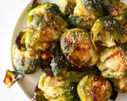 This is an overhead image of a white plate with roasted brussels sprouts on it. The brussels sprouts have parmesan on them. The plate sits on a white counter with crumbs on it.