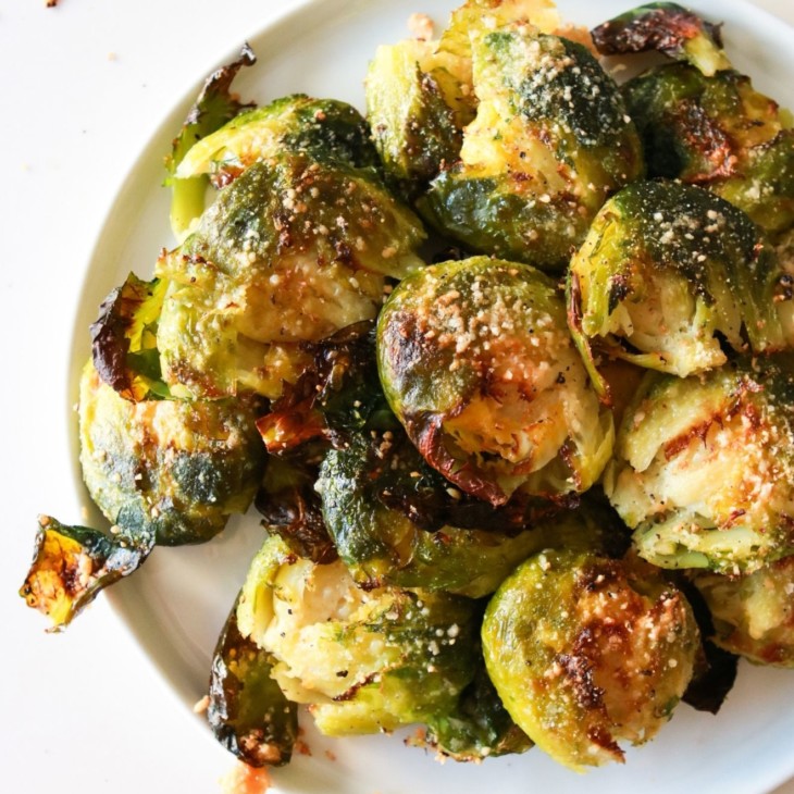 This is an overhead image of a white plate with roasted brussels sprouts on it. The brussels sprouts have parmesan on them. The plate sits on a white counter with crumbs on it.