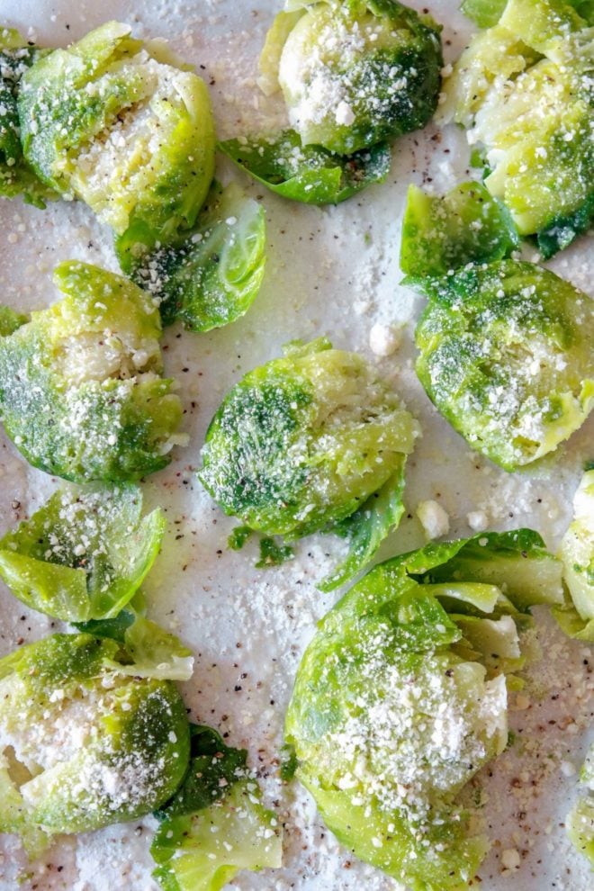 This is an overhead image of brussels sprouts flattened and sprinkled with grated parmesan, salt, and pepper. The brussels sit on a white piece of parchment paper.