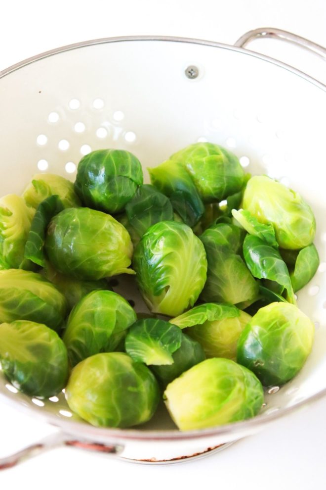 A white colander is filled with bright green boiled brussels sprouts. The colander sits on a white counter.