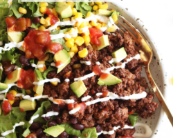 This is an overhead image of a bowl filled with lettuce, ground beef, salsa, avocado, corn, black beans, and sour cream. The bowl sits on a white counter and a gold fork lies partially in the bowl. Text overlay reads "loaded taco salad low carb & gluten free."