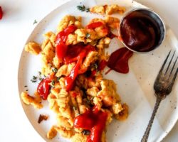 This is an overhead image of a plate with an egg scramble drizzled with ketchup. A fork lays on the right side of the plate along with a small metal cup of ketchup. A cup of orange juice is in the top right corner of the image and some raspberries are scattered on the white counter. A blue tea towel is in the bottom left of the image.