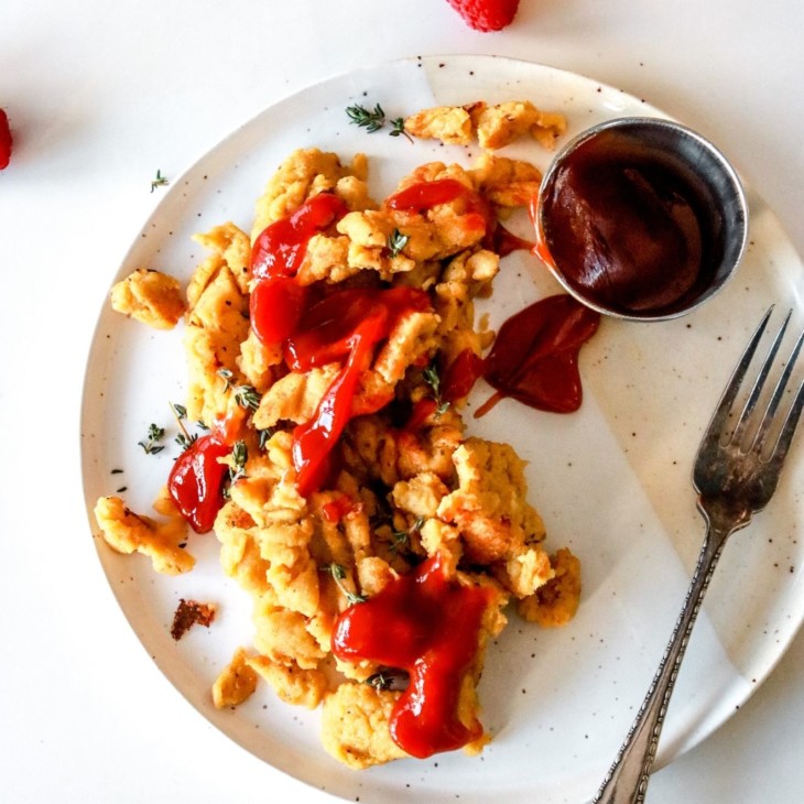 This is an overhead image of a plate with an egg scramble drizzled with ketchup. A fork lays on the right side of the plate along with a small metal cup of ketchup. A cup of orange juice is in the top right corner of the image and some raspberries are scattered on the white counter. A blue tea towel is in the bottom left of the image.