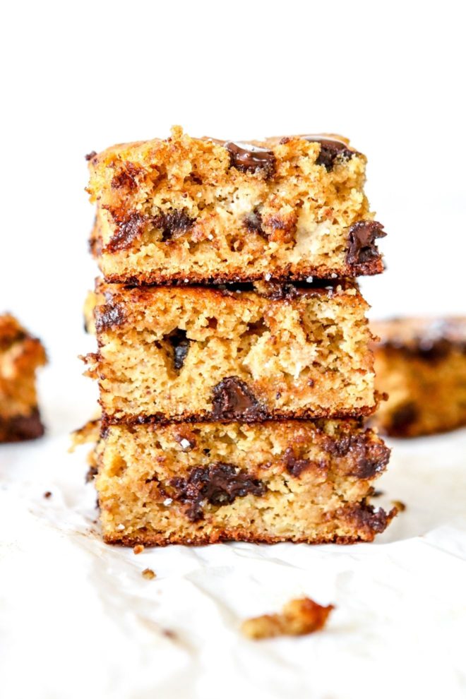 This is a side view of a stack of three chocolate chip banana bars. The bars sit on a white surface and more banana bars are blurred in the background.