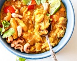 This is an overhead image of a blue bowl filled with jackfruit curry. The curry is topped with lime, cilantro and cashews. A spoon is dipping into the curry and leaning against the side of the bowl. The bowl sits on a white counter with cilantro leaves scattered around it. Another blue bowl filled with curry is in the bottom left of the image.