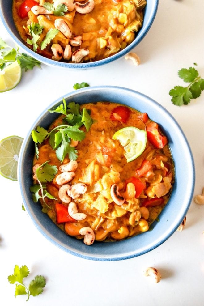 This is an overhead image of blue bowls filled with curry. The curry is garnished with lime wedges, cilantro, and cashews. The bowls sit on a white counter with more cilantro and lime wedges.