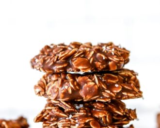 This is a side view of a stack of four chocolate oatmeal cookies. The cookies sit on a white counter with a white background with more cookies blurred in the background.