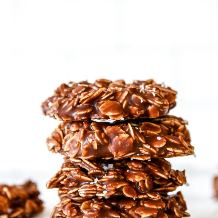 This is a side view of a stack of four chocolate oatmeal cookies. The cookies sit on a white counter with a white background with more cookies blurred in the background.