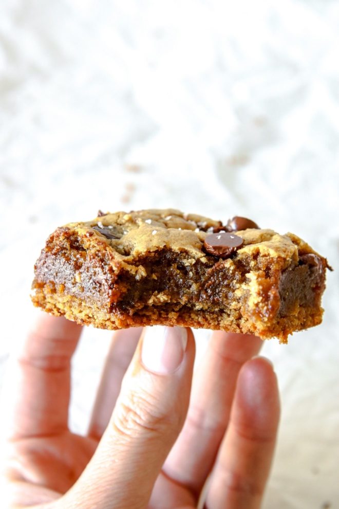 This is a hand holding a chocolate chip tahini bar with a bite taken out of it. There is a crumbled white parchment paper in the background.