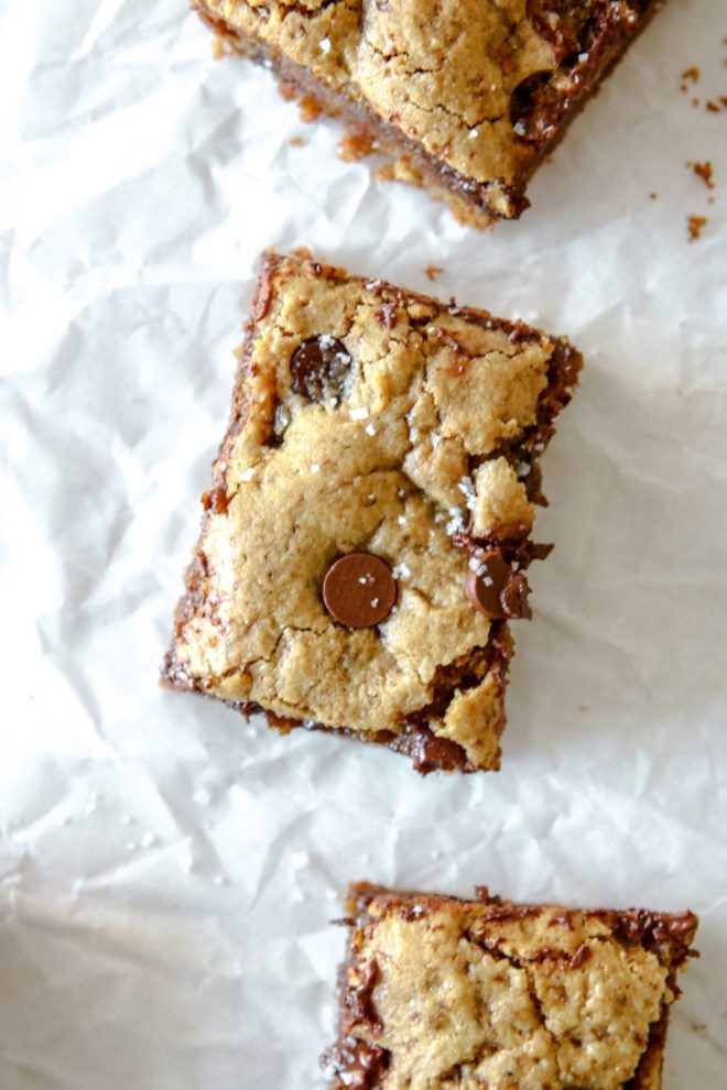 This is an overhead image of three tahini bars with chocolate chips. The bars are in a vertical line with space between them and are on a white piece of parchment paper.