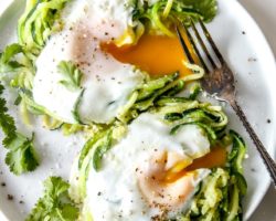 This is an overhead image of a white plate with two mounds of zoodles. In the center of each zoodle is a cooked egg with drippy yellow center. The eggs are cut open and the yellow yolk is dripping out. The egg nests are topped with ground pepper and fresh herbs.