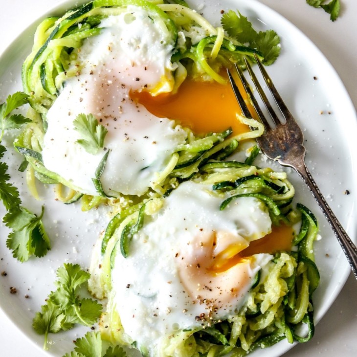 This is an overhead image of a white plate with two mounds of zoodles. In the center of each zoodle is a cooked egg with drippy yellow center. The eggs are cut open and the yellow yolk is dripping out. The egg nests are topped with ground pepper and fresh herbs.