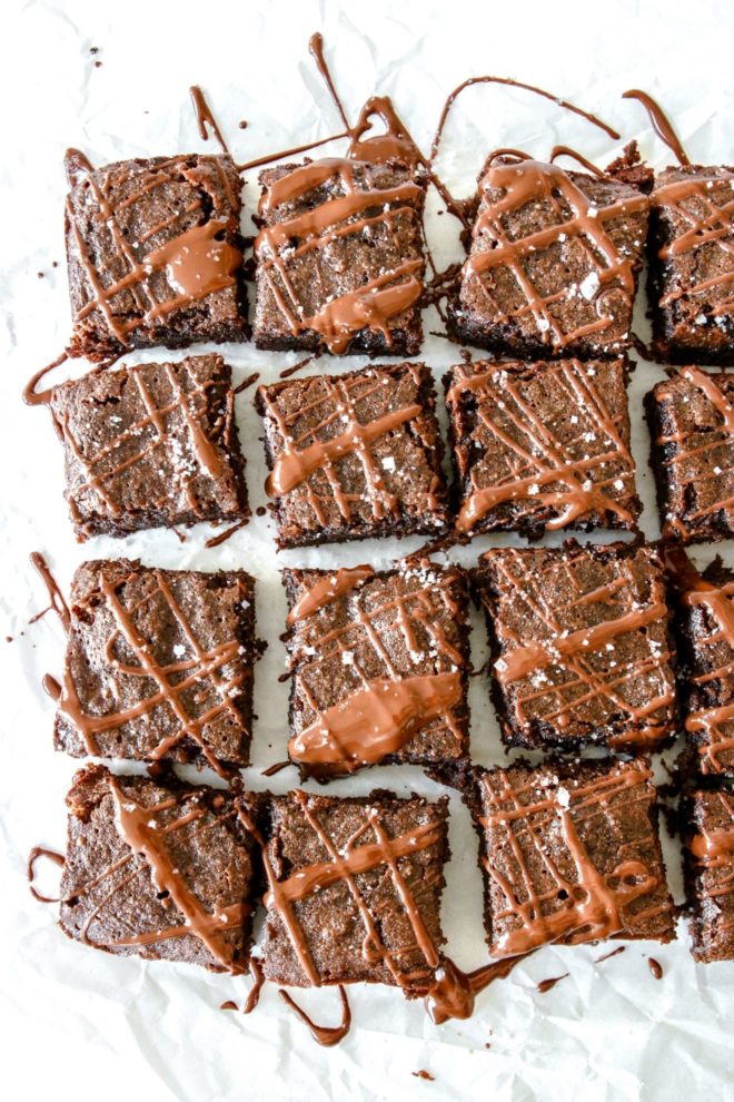 This is an overhead image of 16 brownies on a white piece of parchment paper. The brownies are drizzled with melted chocolate and sprinkled with sea salt.