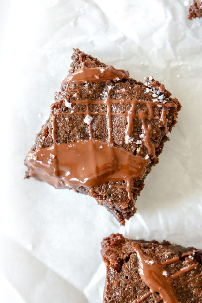 This is an overhead image of a chocolate brownie. The brownie is on a white piece of parchment paper with another brownie in the bottom right corner of the image. The brownies are drizzled with melted chocolate and sprinkled with sea salt.