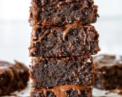 This is a stack of four zucchini brownies with melted chocolate drizzling down the side. The stack sits on a white piece of parchment paper and a white background with more brownies blurred behind the stack.