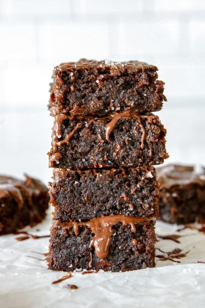 This is a stack of four zucchini brownies with melted chocolate drizzling down the side. The stack sits on a white piece of parchment paper and a white background with more brownies blurred behind the stack.