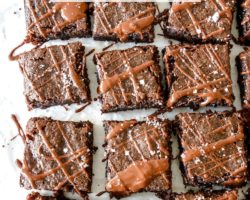 This is an overhead image of 16 brownies on a white piece of parchment paper. The brownies are drizzled with melted chocolate and sprinkled with sea salt. Text overlay reads "deliciously fudgey zucchini brownies."