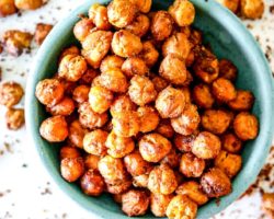 This is an overhead view of a small turquoise bowl filled with crispy chickpeas. The bowl sits on a white counter with more chickpeas scattered around the bowl.