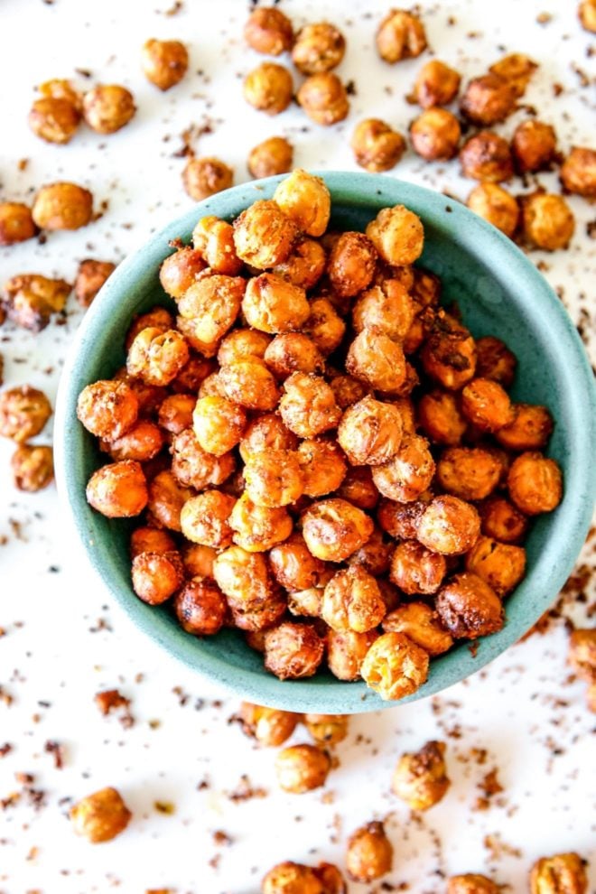 This is an overhead view of a small turquoise bowl filled with crispy chickpeas. The bowl sits on a white counter with more chickpeas scattered around the bowl.