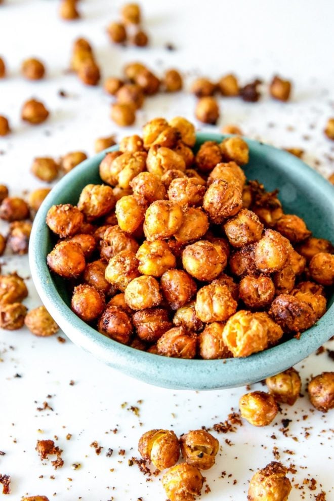 This is a side view of a small turquoise bowl filled with crispy chickpeas. The bowl sits on a white counter with more chickpeas scattered around the bowl.