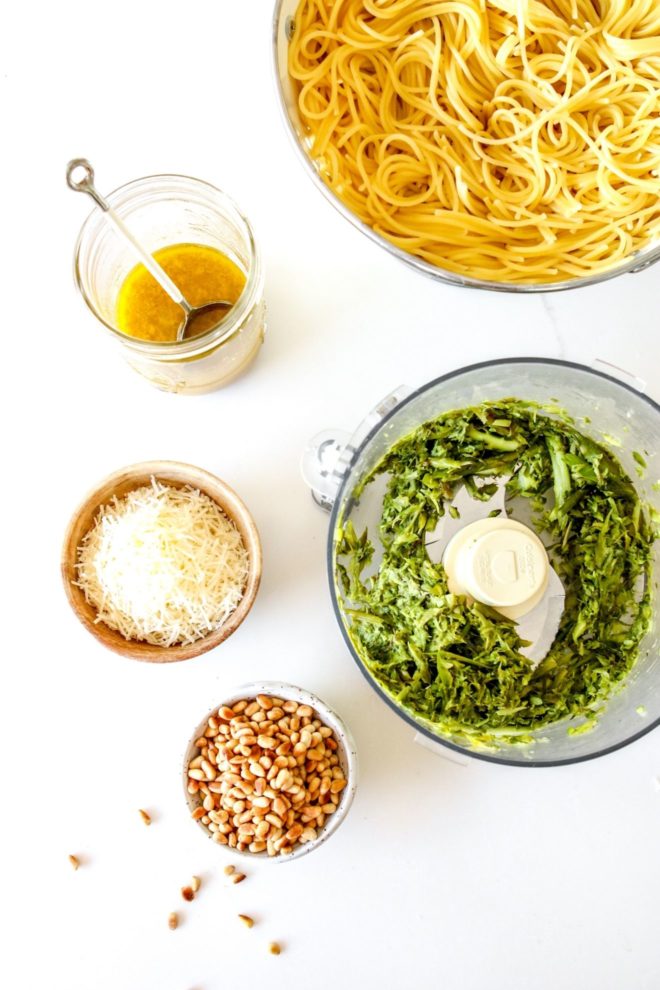 This is an overhead view of cooked spaghetti in a colander, chopped asparagus in a food processor, shredded parmesan in a bowl, toasted pine nuts in a bowl, and lemon dressing in a mason jar. The ingredients sit on a white counter.