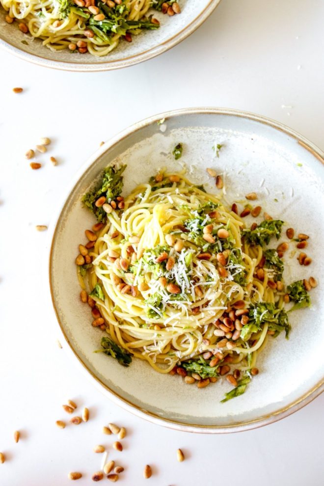 This is an overhead view of a shallow bowl sitting on a white counter. In the bowl is spaghetti topped with shredded asparagus and toasted pine nuts.