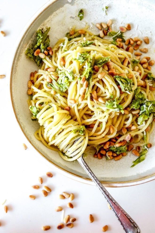 This is an overhead view of a shallow bowl with spaghetti topped with shredded asparagus and toasted pine nuts. A fork has spaghetti twirled around the fork and leaning against the side of the bowl.