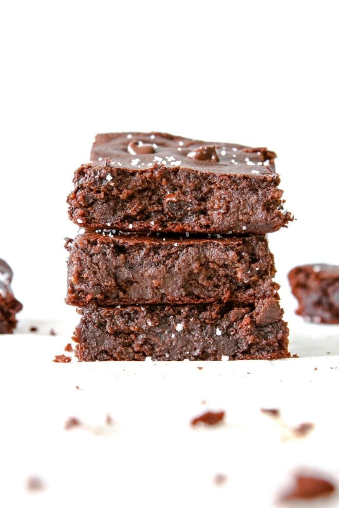 This is a side view of a stack of three chickpea brownies. The brownies sit on a white surface with a white background and are sprinkled with salt. More gluten free brownies are off to the side and blurred in the background. 