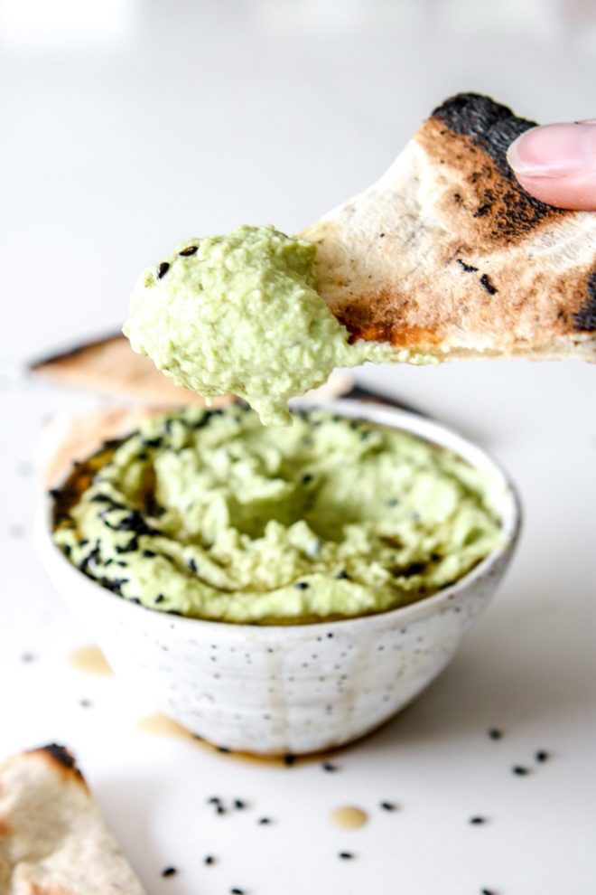 This is a side image of a white speckled bowl filled with light green edamame hummus. A hand is holding a piece of pita that has been dipped in the hummus and holding it up above the bowl.