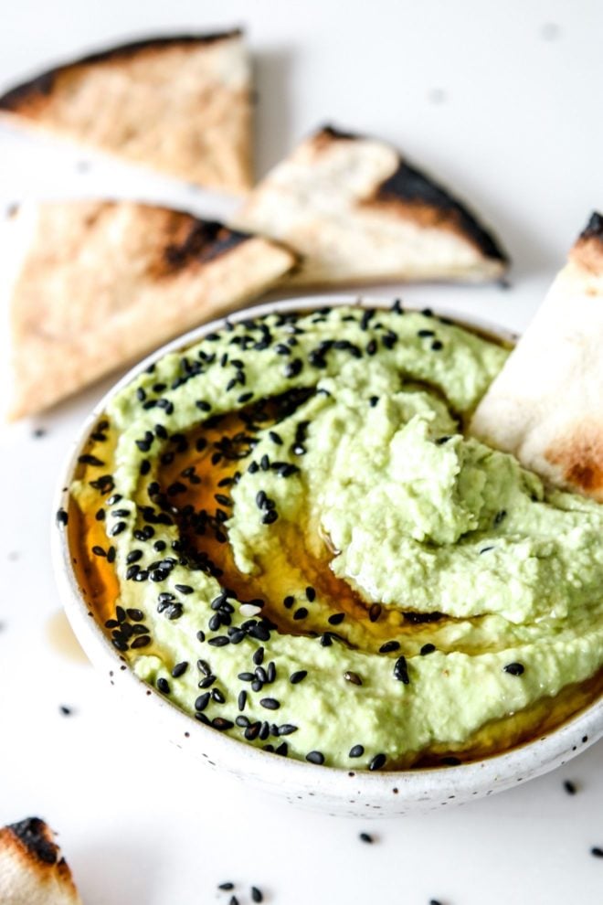 This is a side image of a white bowl sitting on a white counter with triangle pieces of pita around it. Inside the bowl is a light green edamame hummus with black sesame seeds and oil drizzled on top. A pita is dipped on the side.