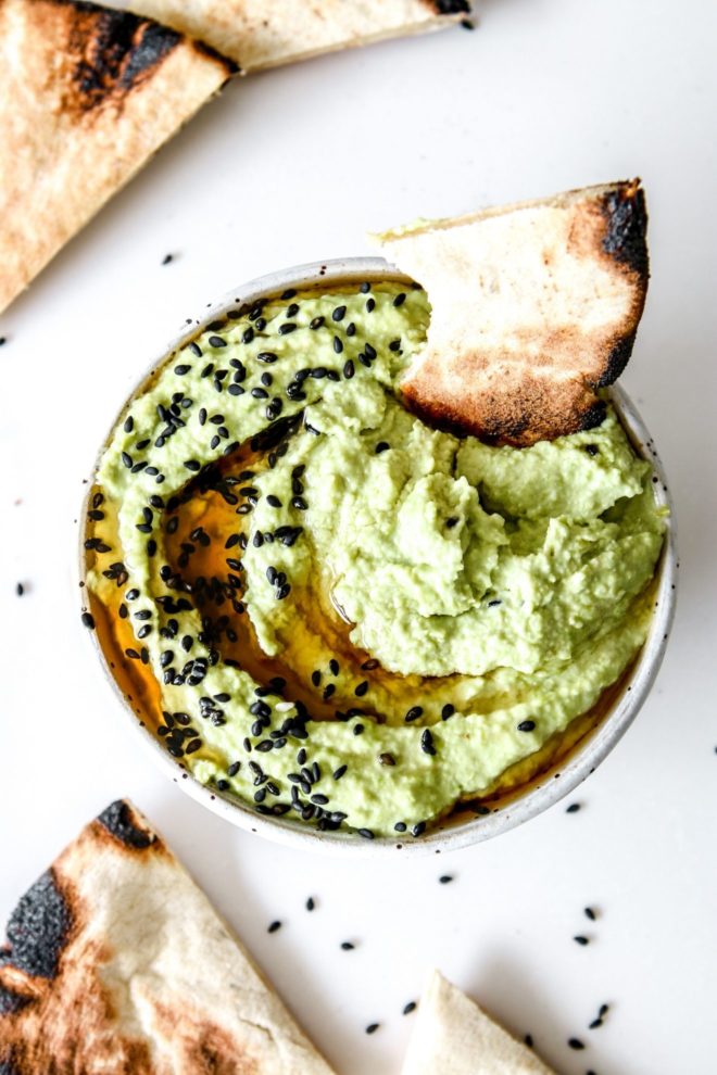This is an overhead image of a white bowl with light green edamame hummus in it. The hummus has black sesame seeds and oil drizzled on top. A piece of pita bread is dipping into the hummus toward the top of the bowl. The bowl sits on a white counter with more piece of pita bread and black sesames on the counter.