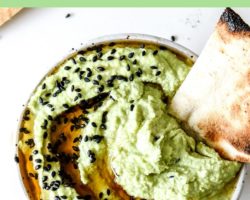 This is an overhead image of a white bowl with light green edamame hummus in it. The hummus has black sesame seeds and oil drizzled on top. A piece of pita bread is dipping into the hummus toward the top of the bowl. The bowl sits on a white counter with more piece of pita bread and black sesames on the counter. Text overlay reads "edamame hummus."