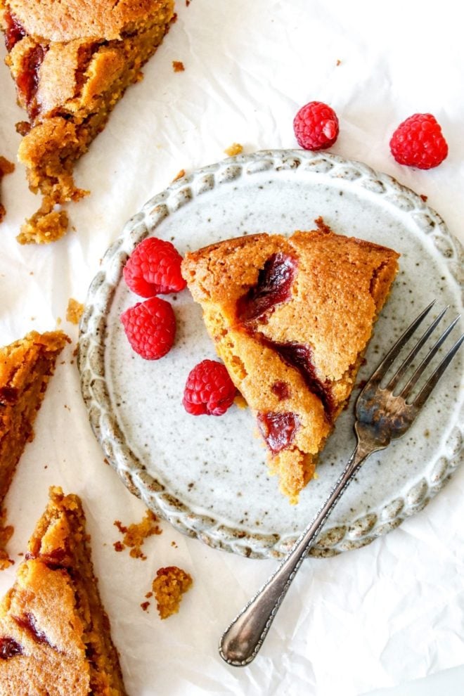 This is an overhead image of a slice of lemon cake with a raspberry jam swirl on top. The slice sits on a small speckled plate with an antique fork and fresh raspberries. The plate sits on a white piece of parchment paper with more slices of cake and fresh raspberries.