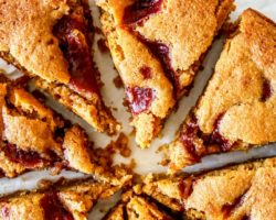This is an overhead close-up image of a lemon cake with raspberry jam swirl on top. The cake is cut into 8 triangles. The cake sits on a white surface with fresh raspberries on the side.