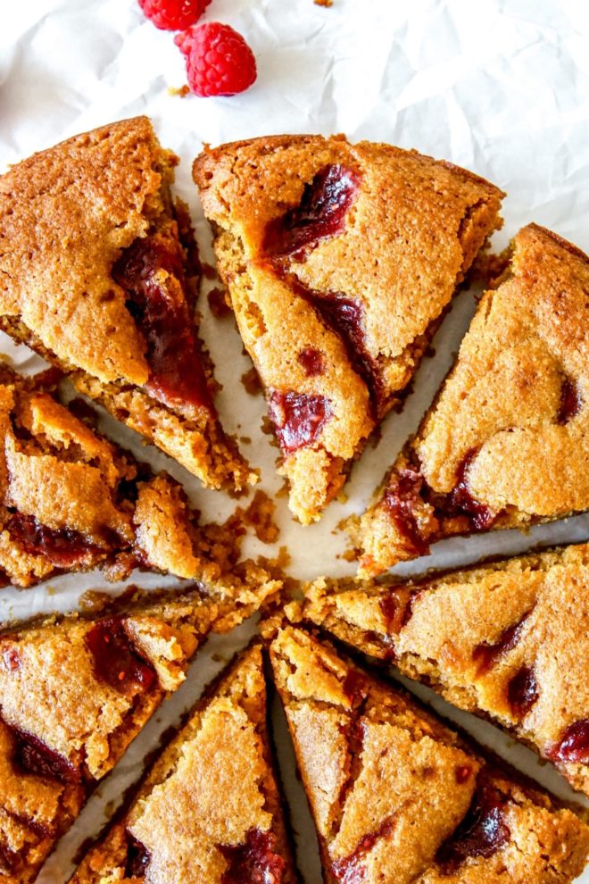 This is an overhead close-up image of a lemon cake with raspberry jam swirl on top. The cake is cut into 8 triangles. The cake sits on a white surface with fresh raspberries on the side.