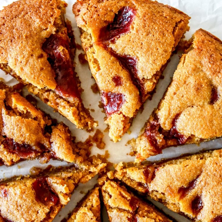 This is an overhead close-up image of a lemon cake with raspberry jam swirl on top. The cake is cut into 8 triangles. The cake sits on a white surface with fresh raspberries on the side.