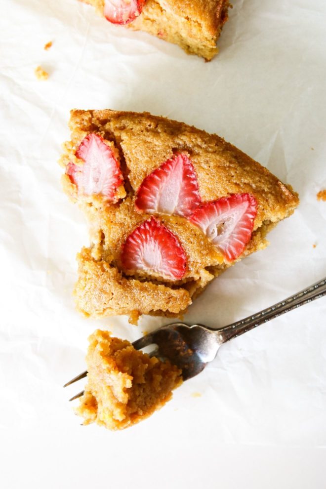 This is an overhead view of a slice of lemon cake topped with sliced strawberries. A fork is taking a piece off the cake. The cake slice is on a white piece of parchment paper.