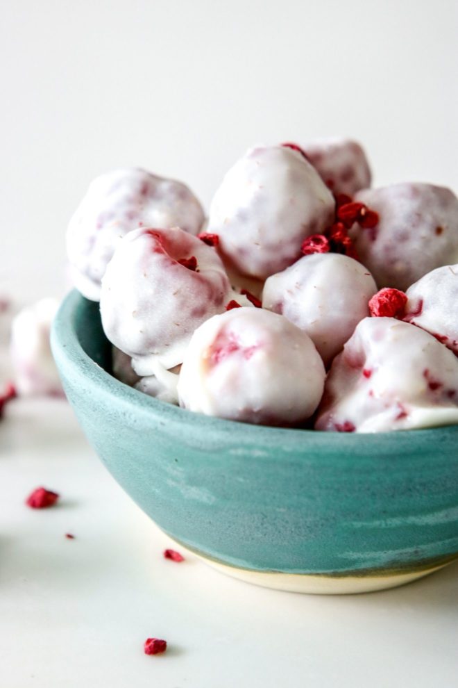 This is a side view of a small teal bowl with frozen raspberries coated in white chocolate. The bowl sits on a white counter with more frozen raspberries on the counter next to the bowl.