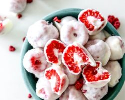 This is an overhead image of a small teal bowl with frozen raspberries coated in white chocolate. The bowl sits on a white counter with more frozen raspberries on the counter next to the bowl.