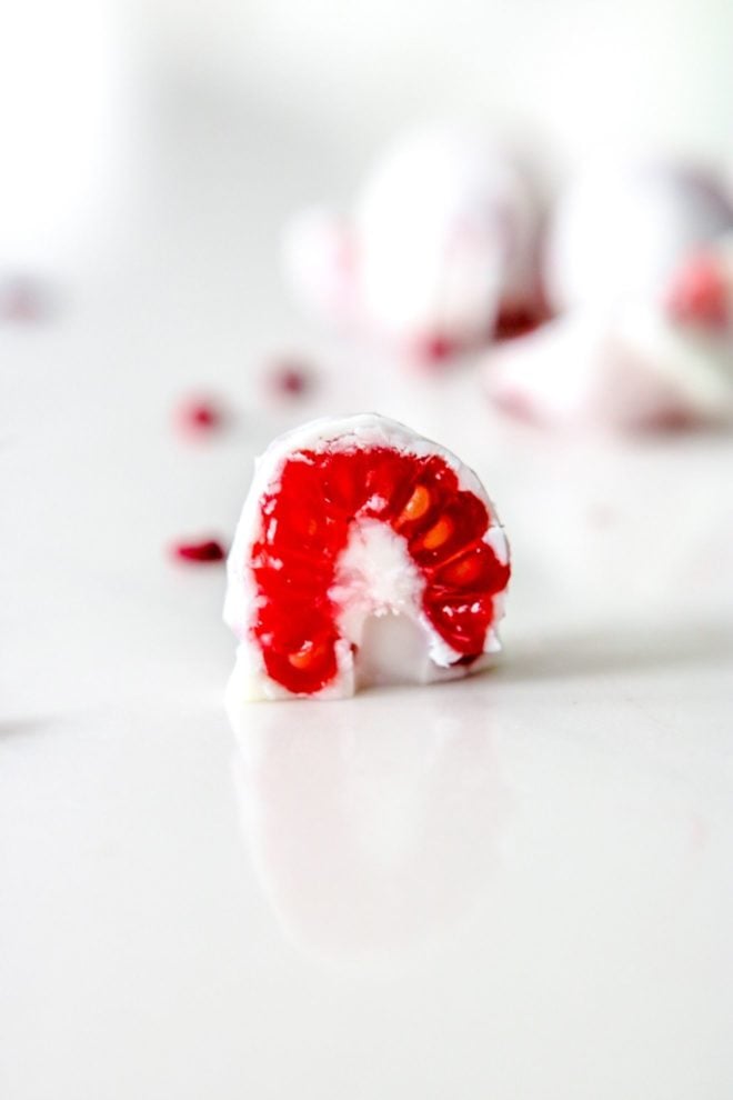 This is a side image of a raspberry coated in white chocolate cut in half. The raspberry sits on a white counter. More white chocolate coated raspberries are blurred in the background.