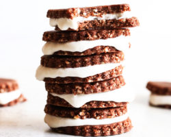 This is a side view of a stack of homemade chocolate sandwich cookies. The cookie sandwiches have a vanilla icing inside. The top cookie has a bite taken out. More cookies are blurred in the background.