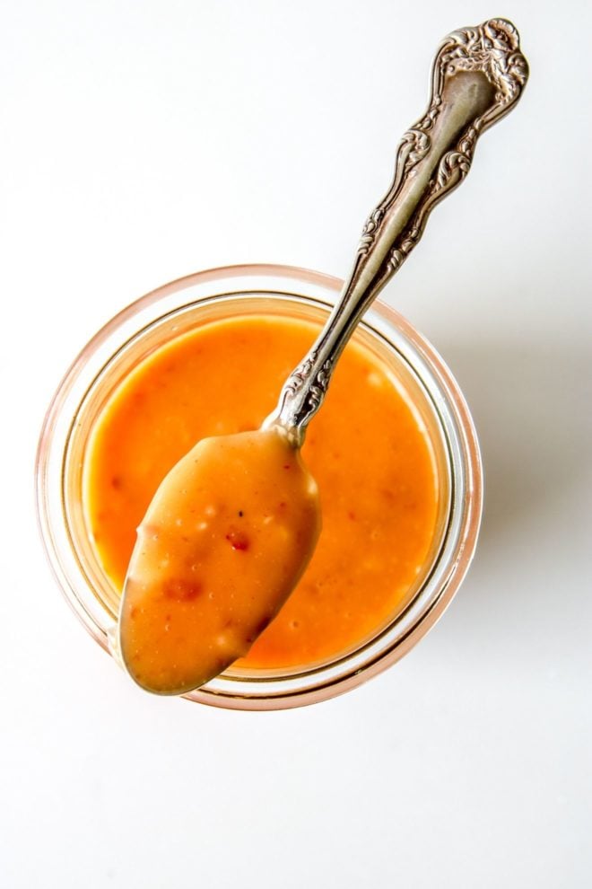 This is an overhead image of a glass jar with orange bang bang sauce in it. An antique spoon is leaning on top of the jar with a scoop of sauce in it. The jar is sitting on a white counter.
