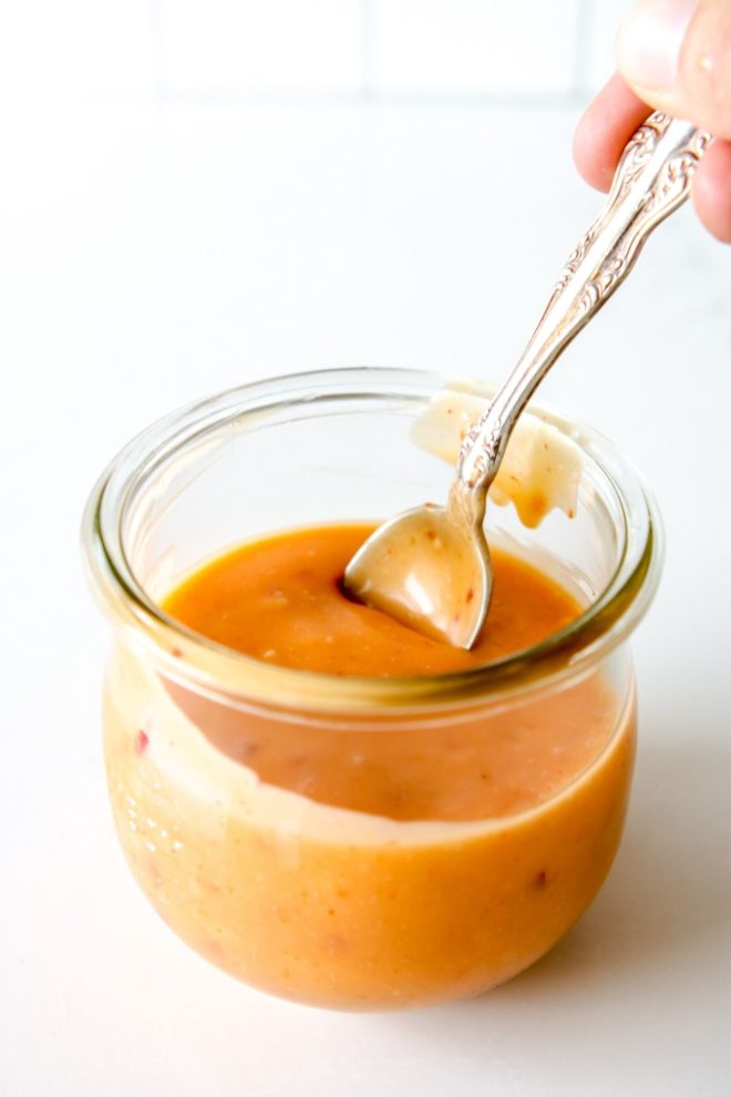 This is an overhead side-view of a glass jar filled with orange bang bang sauce. A spoon is scooping and drizzling the sauce back into the jar. The jar sits on a white counter with a white background.