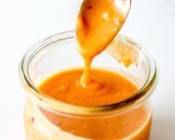 This is an overhead side-view of a glass jar filled with orange bang bang sauce. A spoon is scooping and drizzling the sauce back into the jar. The jar sits on a white counter with a white background.