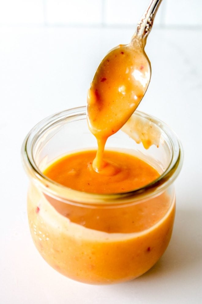 This is an overhead side-view of a glass jar filled with orange bang bang sauce. A spoon is scooping and drizzling the sauce back into the jar. The jar sits on a white counter with a white background.