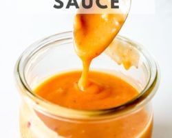 This is an overhead side-view of a glass jar filled with orange bang bang sauce. A spoon is scooping and drizzling the sauce back into the jar. The jar sits on a white counter with a white background. Text overlay reads "easy 5 minute bang bang sauce."