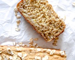 This is an overhead image of a slice of banana bread on a white piece of parchment paper. There are oats sprinkled around the bread. The full loaf is blurred at the bottom of this image.