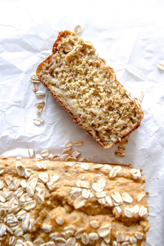 This is an overhead image of a slice of banana bread on a white piece of parchment paper. There are oats sprinkled around the bread. The full loaf is blurred at the bottom of this image.